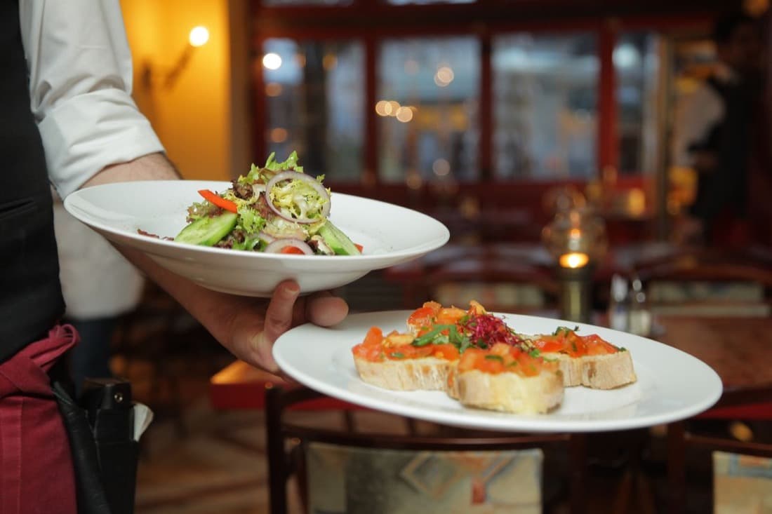 A waiter carrying two plated dishes through a restaurant dining room